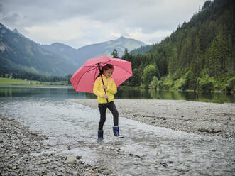 Verspieltes Mädchen mit Regenschirm, das am Seeufer auf dem Wasser läuft - DIKF00652