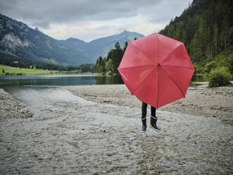 Girl covering face behind red umbrella standing on water at lakeshore - DIKF00656