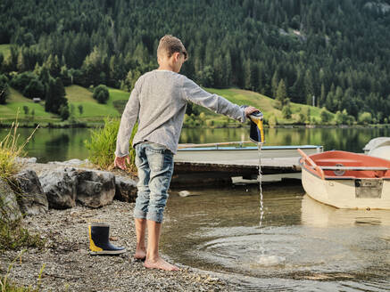 Junge entfernt Wasser aus Gummistiefel am Seeufer - DIKF00660