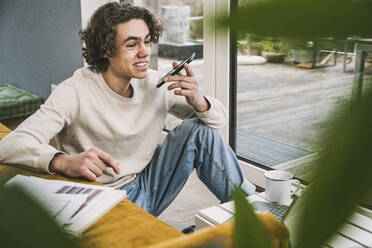 Lächelnder junger Mann, der mit seinem Smartphone über einen Lautsprecher im Wohnzimmer spricht - UUF25541