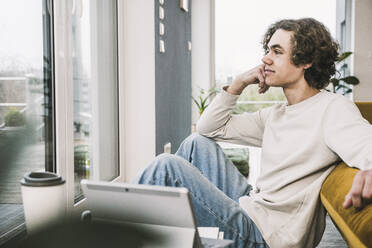 Young man sitting with hand on chin by on sofa in living room - UUF25543