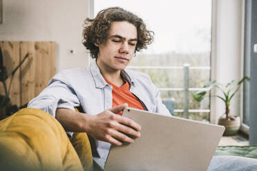 Young man with laptop sitting on sofa in living room at home - UUF25545