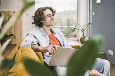 Thoughtful smiling man with laptop sitting on sofa in living room at home - UUF25546