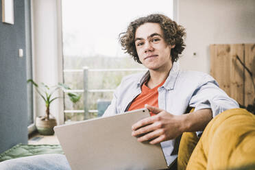 Smiling man with laptop sitting on sofa in living room - UUF25548