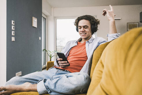 Happy man listening music through wireless headphones using smart phone on sofa at home - UUF25551