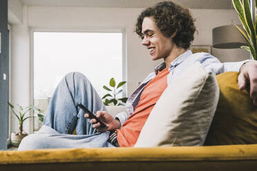 Happy young man surfing net through mobile phone sitting on sofa at home - UUF25553