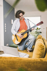 Young man playing guitar on sofa in living room at home - UUF25558