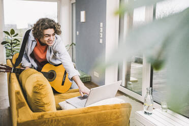 Smiling young man holding guitar using laptop on sofa at home - UUF25559