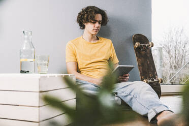 Junger Mann mit Tablet-PC, der zu Hause neben einem Skateboard sitzt - UUF25561