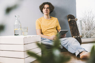 Junger Mann mit Tablet-PC, der zu Hause neben einem Skateboard sitzt - UUF25563