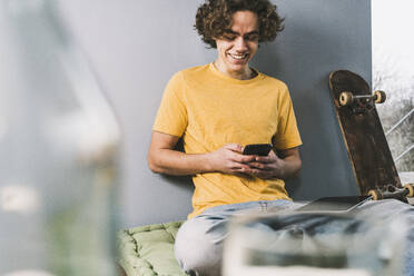 Glücklicher Mann, der zu Hause auf dem Skateboard eine Textnachricht über sein Smartphone verschickt - UUF25565
