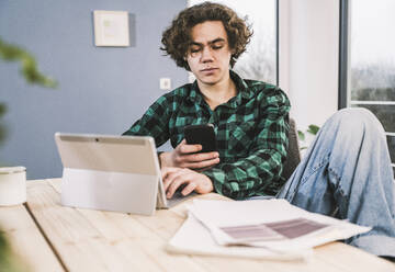 Young student sitting with tablet PC using mobile phone at table in living room - UUF25570