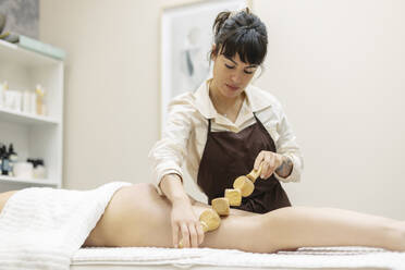 Therapist massaging woman's thigh with wooden roller at aesthetic clinic - MTBF01174