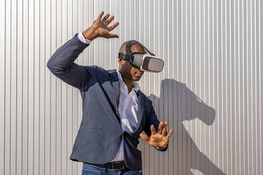 Geschäftsmann mit Virtual-Reality-Simulator gestikuliert vor einer Wand an einem sonnigen Tag - DLTSF02653