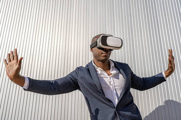 Geschäftsmann, der einen Virtual-Reality-Simulator trägt, steht mit ausgestreckten Armen vor einer Wand an einem sonnigen Tag - DLTSF02656