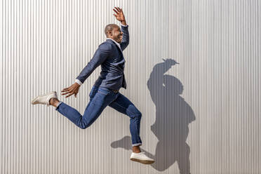 Happy businessman with hand raised jumping by wall on sunny day - DLTSF02660