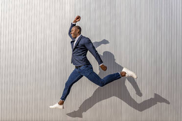 Happy businessman jumping in front of wall on sunny day - DLTSF02662