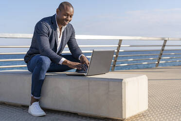 Lächelnder Geschäftsmann mit Laptop auf einer Bank an der Promenade sitzend - DLTSF02667