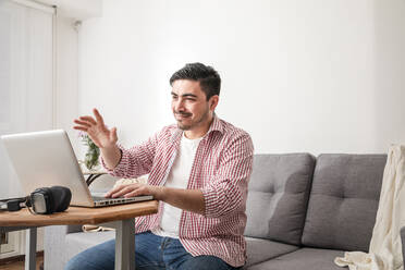 Lächelnder Freiberufler mit winkender Hand bei einem Videogespräch über einen Laptop zu Hause - FLMF00788