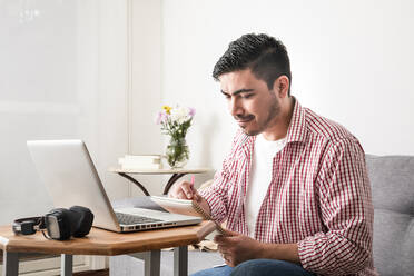 Lächelnder Mann, der im Wohnzimmer am Laptop in einen Notizblock schreibt - FLMF00789