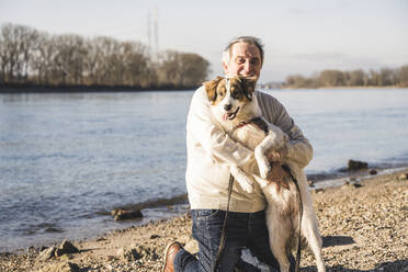 Lächelnder älterer Mann umarmt Hund am Strand an einem sonnigen Tag - UUF25592