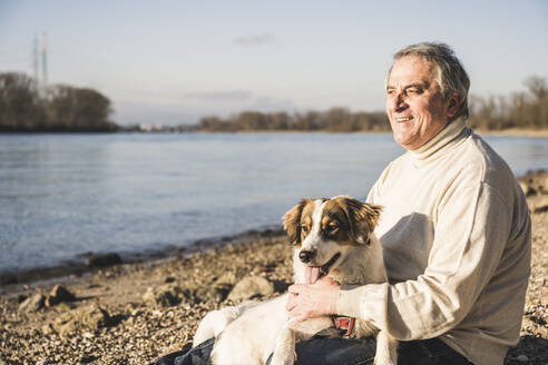 Glücklicher Mann mit Hund am Strand sitzend an einem sonnigen Tag - UUF25594