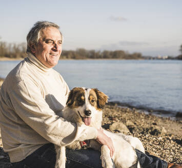 Lächelnder älterer Mann mit Hund am Strand sitzend an einem sonnigen Tag - UUF25595