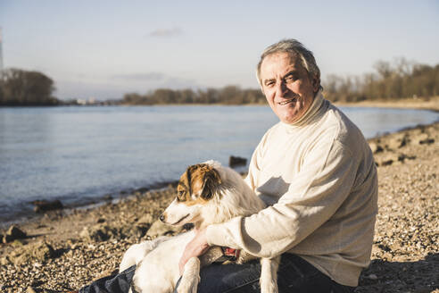 Glücklicher älterer Mann mit Hund am Strand sitzend an einem sonnigen Tag - UUF25596