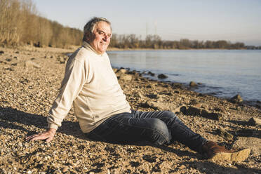 Lächelnder Mann sitzt am Strand an einem sonnigen Tag - UUF25604