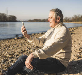 Glücklicher älterer Mann führt einen Videoanruf über ein Mobiltelefon am Strand an einem sonnigen Tag durch - UUF25605