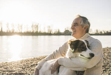 Lächelnder älterer Mann mit Hund am Strand an einem sonnigen Tag - UUF25613