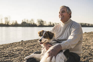 Glücklicher Mann mit Hund am Strand sitzend an einem sonnigen Tag - UUF25615