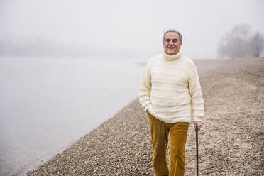 Glücklicher Mann in warmer Kleidung, der mit der Hand in der Tasche am Strand spazieren geht - UUF25623