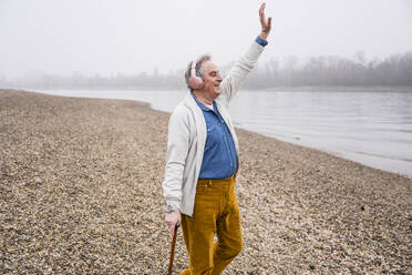 Glücklicher älterer Mann mit erhobener Hand hört Musik über Kopfhörer am Strand - UUF25634