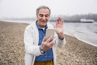 Glücklicher älterer Mann, der einen Videoanruf über sein Smartphone tätigt und mit der Hand am Strand winkt - UUF25643