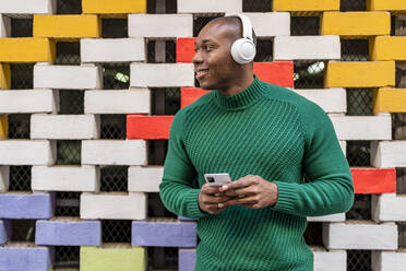 Lächelnder Mann hört Musik über drahtlose Kopfhörer vor einer mehrfarbigen Wand - DLTSF02706