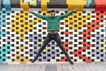 Happy man with arms outstretched jumping in front of multi colored wall - DLTSF02711