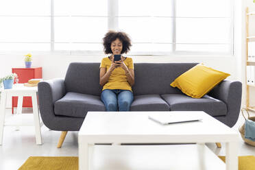 Smiling young woman using smart phone sitting on sofa in living room at home - XLGF02689