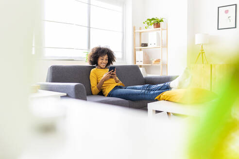 Smiling woman with smart phone sitting on sofa in living room at home - XLGF02696