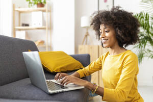 Happy young woman using laptop by sofa at home - XLGF02703