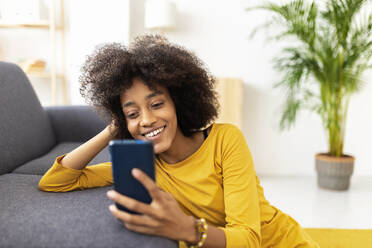 Smiling woman using smart phone leaning on sofa at home - XLGF02705