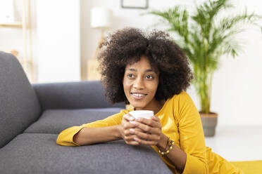 Thoughtful young woman with coffee cup sitting by sofa at home - XLGF02707
