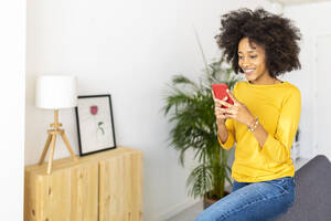 Glückliche Frau, die auf dem Sofa zu Hause sitzt und über ihr Handy SMS schreibt - XLGF02713