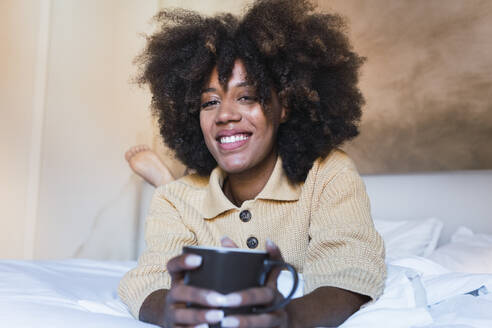 Lächelnde Frau mit Kaffeetasse auf dem Bett im Schlafzimmer liegend - PNAF03114