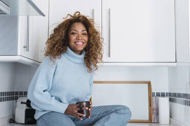 Lächelnde Frau mit Kaffeetasse in der Küche zu Hause sitzend - PNAF03119