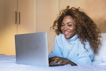 Happy woman using laptop lying on bed in bedroom - PNAF03121