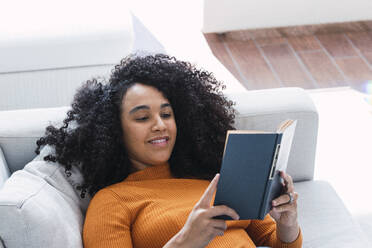 Woman reading book lying on sofa in living room - PNAF03157