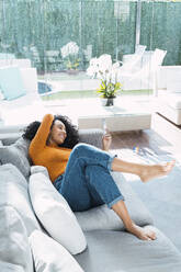 Woman using smart phone lying on sofa in living room at home - PNAF03191