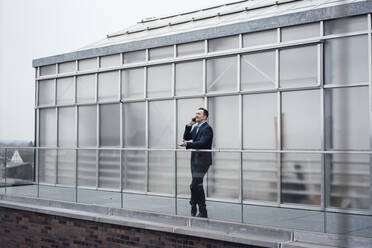 Geschäftsmann, der außerhalb eines Gewächshauses an einem Glasgeländer mit seinem Smartphone telefoniert - MOEF03986