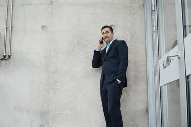 Geschäftsmann mit Hand in der Tasche, der an einer Wand im Gewächshaus mit seinem Smartphone telefoniert - MOEF04012